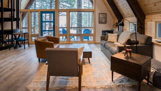 A cozy loft-style room with a high ceiling, large windows, and a view of the forest. - Salted Roots Blue Door A-Frame Cabin (Seward)
