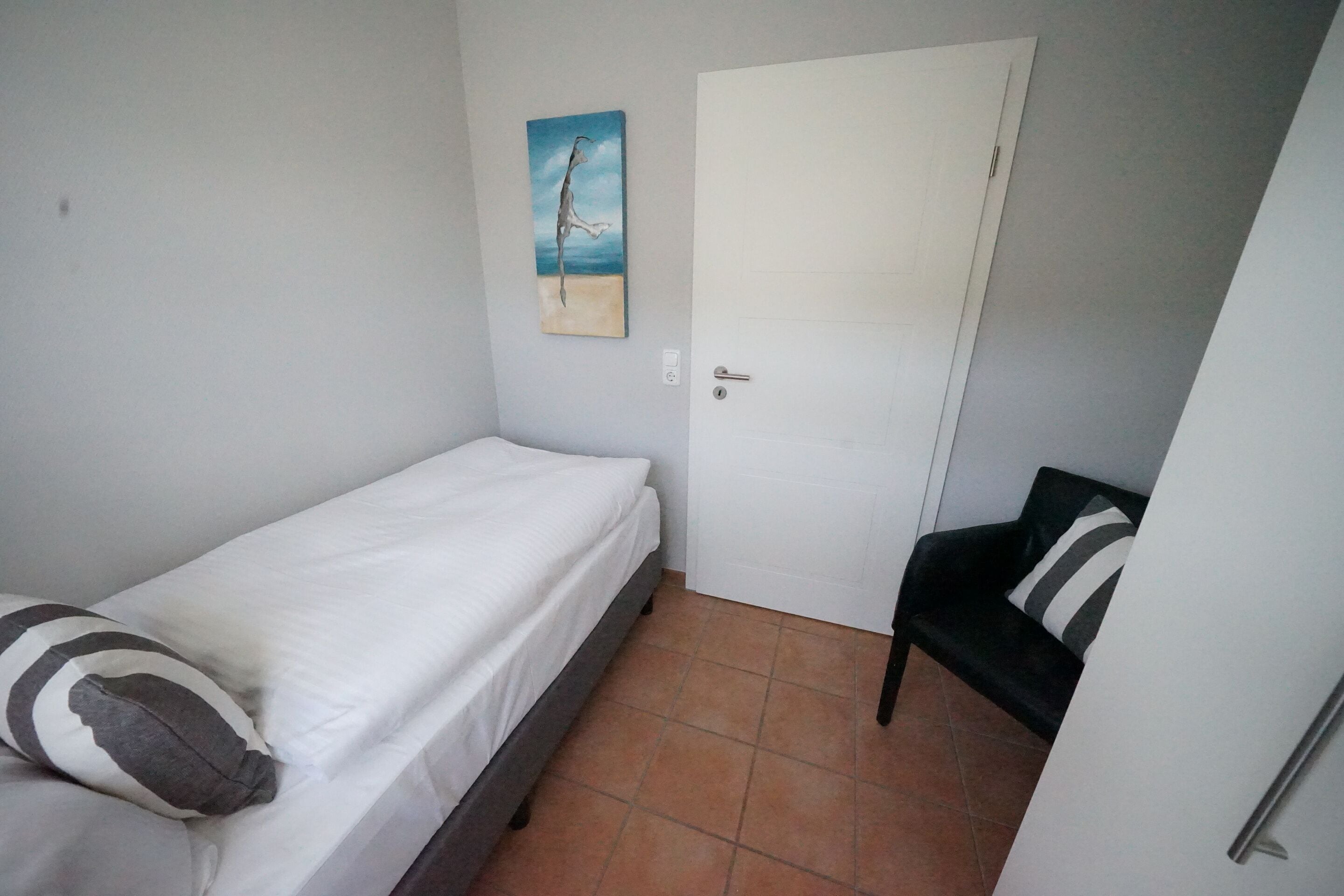 Quarto pequeno com uma cama de solteiro, parede branca lisa, uma porta e uma cadeira com almofadas listradas.