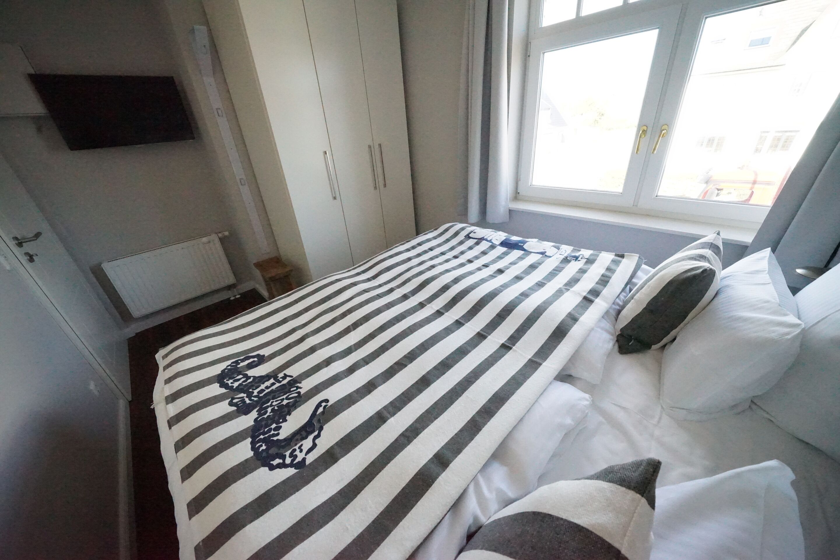 A bedroom with a striped bedspread, white bedding, a flat-screen TV mounted on the wall, and a window with curtains.