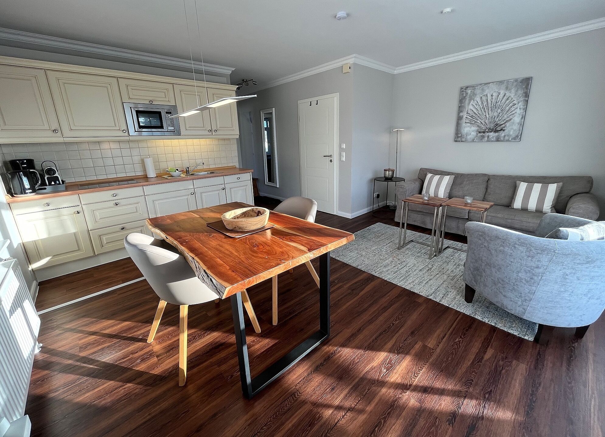 A modern kitchen with white cabinets, a wooden dining table, and a grey sofa in the living area.