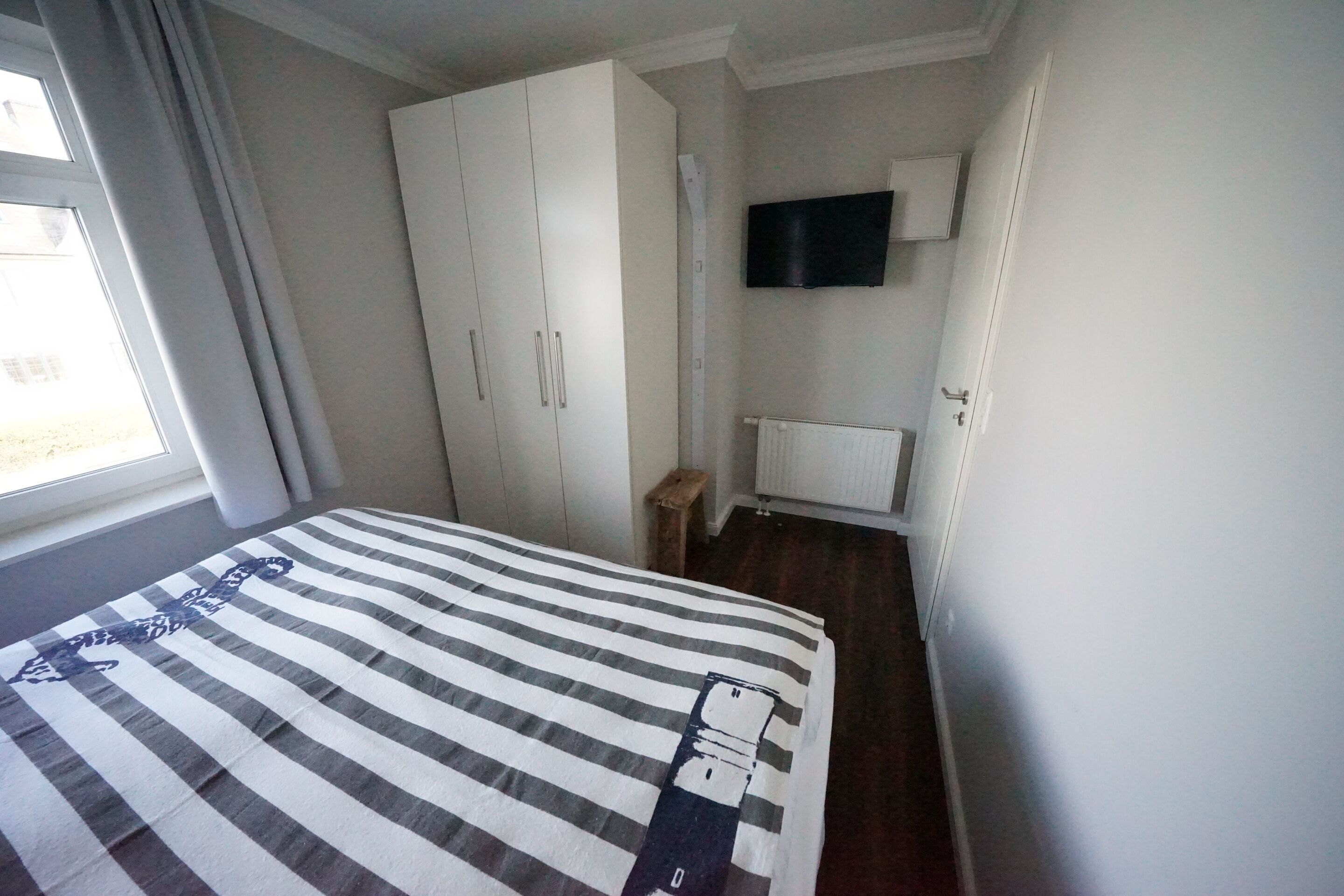 A bedroom with a bed, a wardrobe, a television mounted on the wall, and a window with curtains.