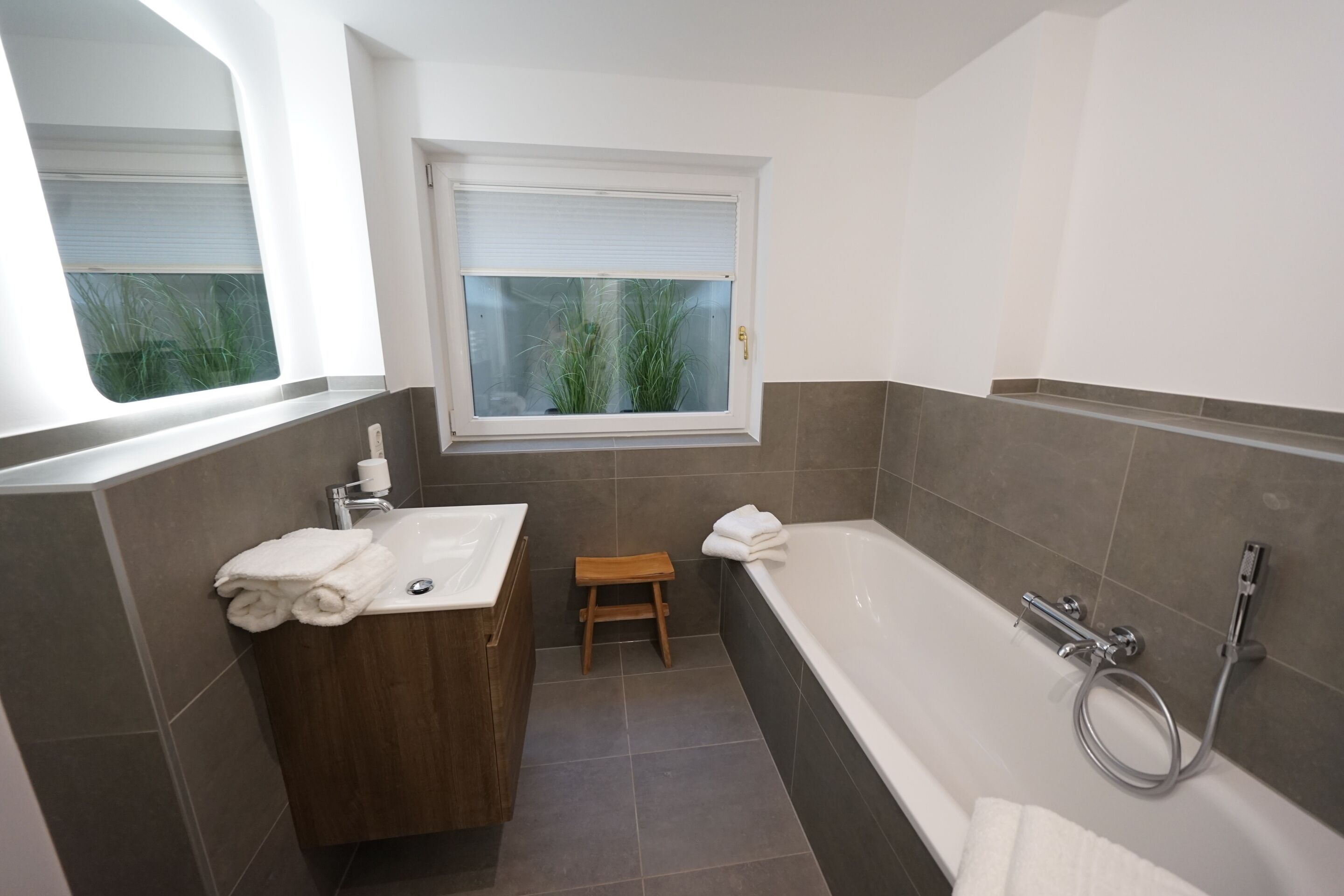 A modern bathroom with a wooden vanity, a bathtub, and a window with blinds.