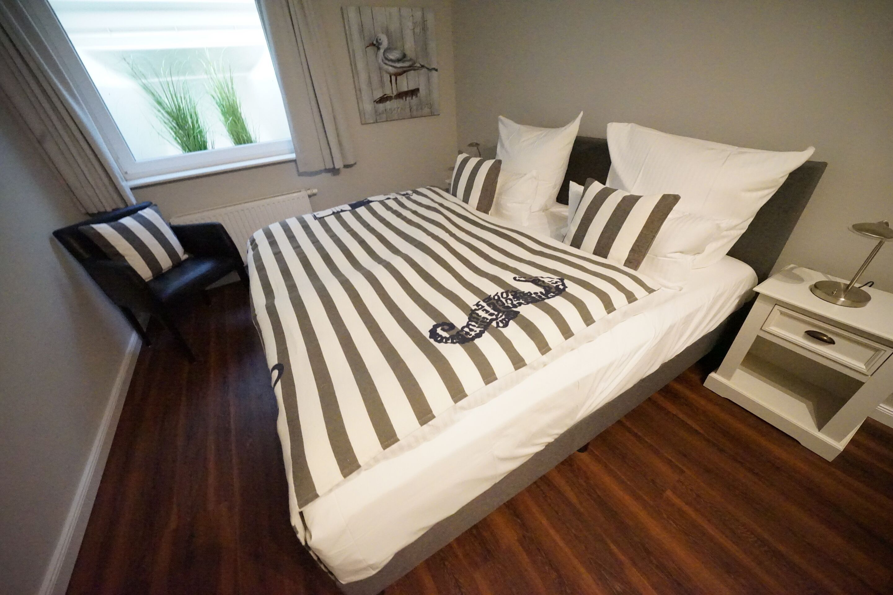 Uma cama bem arrumada com roupa de cama listrada, uma poltrona de couro preto, uma mesa de cabeceira com abajur e uma janela com cortinas.