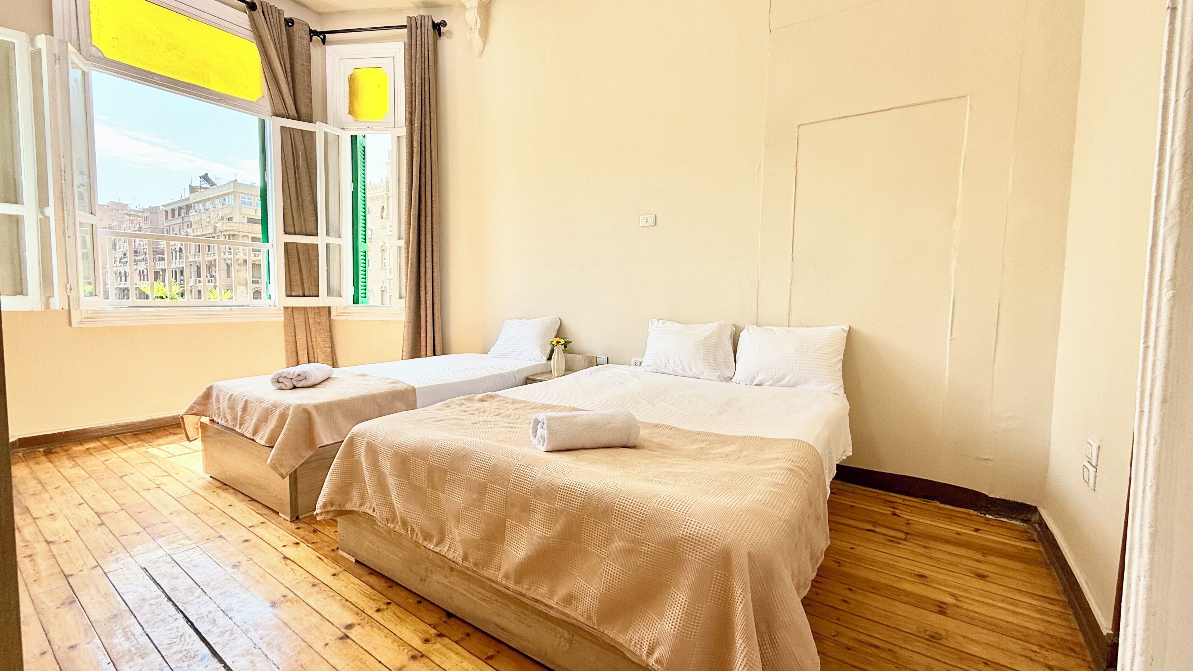 A hotel room with two beds, wooden flooring, a window with curtains, and a view of buildings outside.