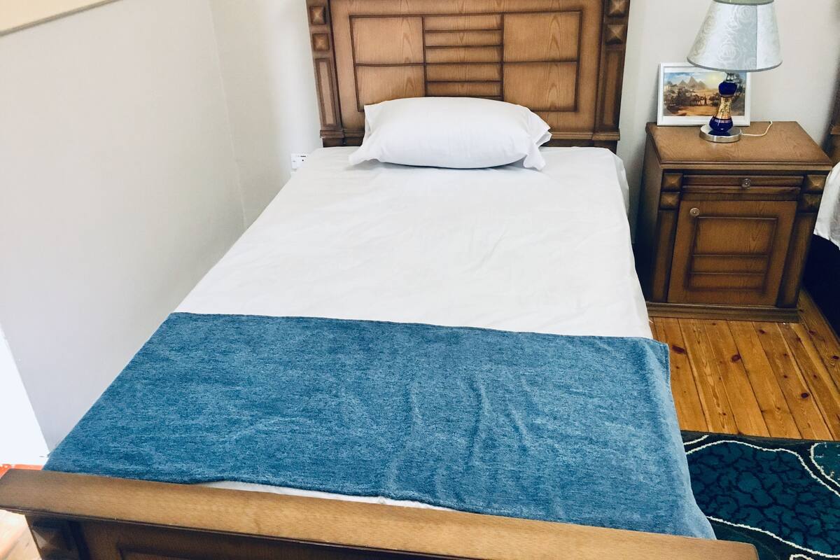 Una cama individual con cabecera de madera, almohada blanca, manta azul, mesita de noche con lámpara y un cuadro en la pared.