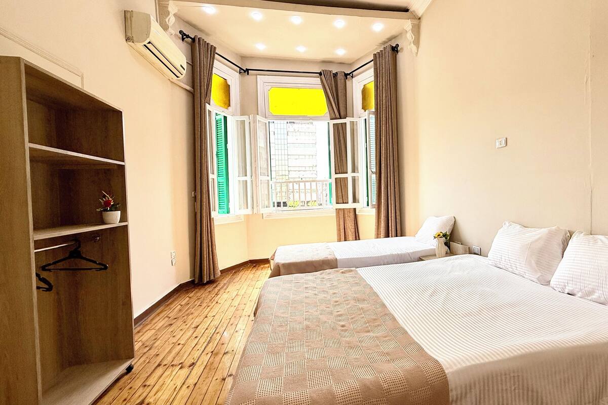 Un dormitorio con dos camas, un armario de madera, un ventanal con cortinas y un balcón pequeño.