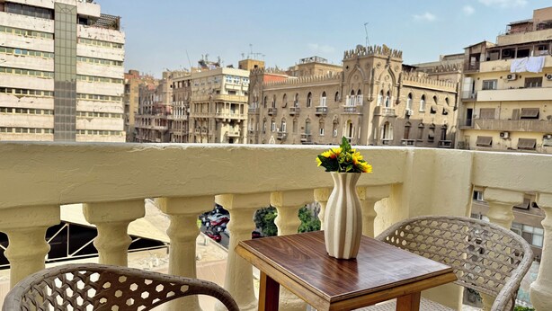 A balcony with a table and chairs, a vase with flowers, and a view of historic buildings. - Bella Casa Hostel (Cairo)