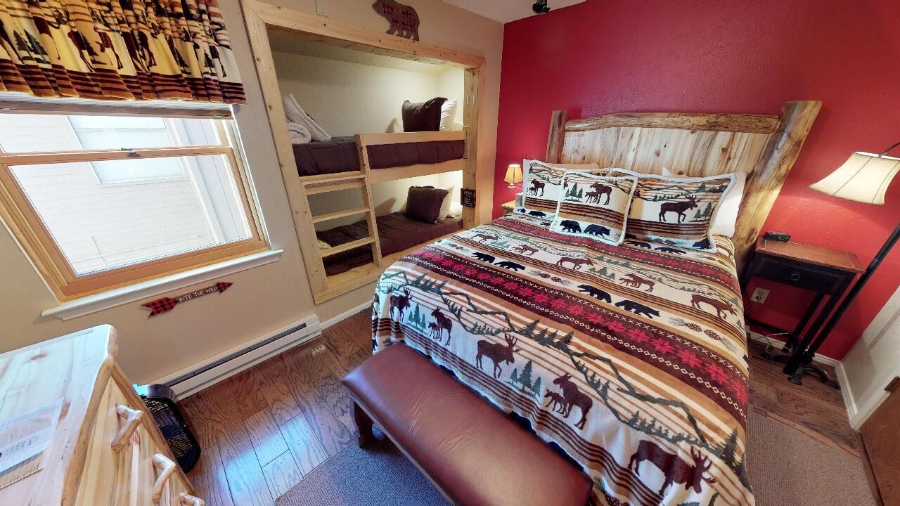 Quarto com beliche, um banco, janela com persianas, parede vermelha e colcha estampada.