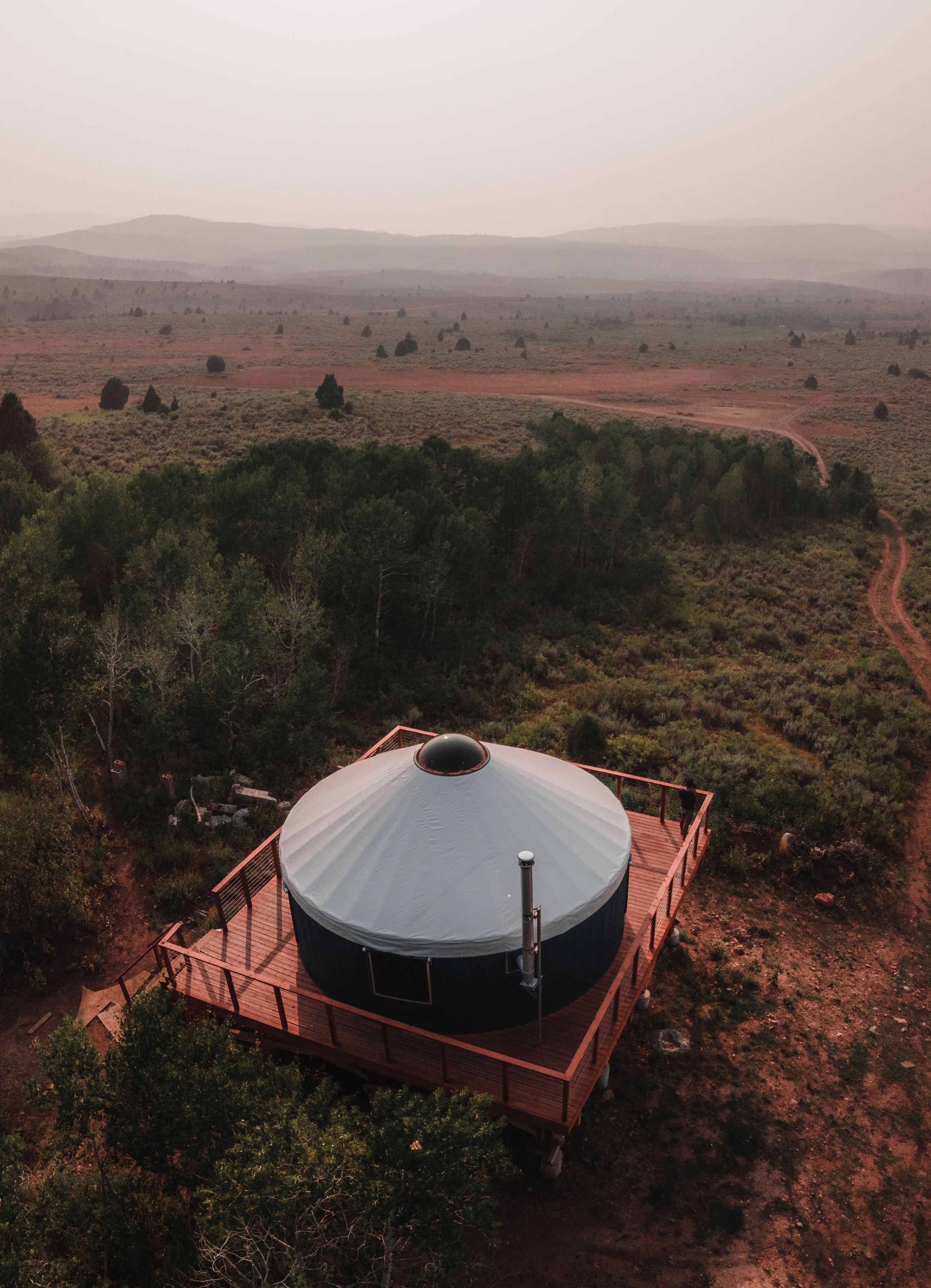 Une grande structure en forme de dôme, surmontée d’un toit plat, entourée d’une terrasse en bois et située dans un paysage naturel caractérisé par une végétation clairsemée et des collines lointaines.