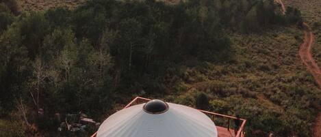 Une grande structure en forme de dôme, surmontée d’un toit plat, entourée d’une terrasse en bois et située dans un paysage naturel caractérisé par une végétation clairsemée et des collines lointaines.