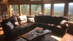 A cozy living room with a large L-shaped leather couch, a coffee table, and a view of a forested landscape through large windows. - Luxury MTN House *Breathtaking Views* Hot Tub* 2 Outdoor Fireplaces *Fire Pit (Blue Ridge)