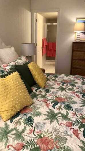 Une chambre avec une couvre-lit à motifs floraux, une commode en bois et une porte donnant sur une salle de bain où est accrochée une serviette rouge.