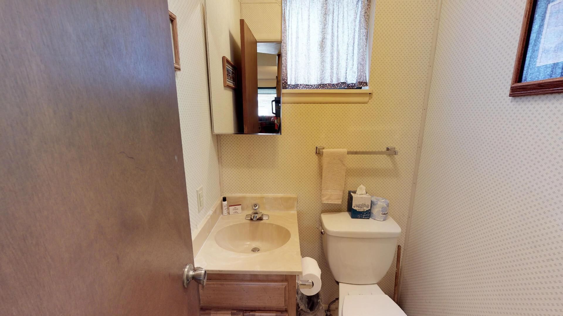 A small bathroom with a wooden cabinet, a sink, a toilet, and a window with a lace curtain.