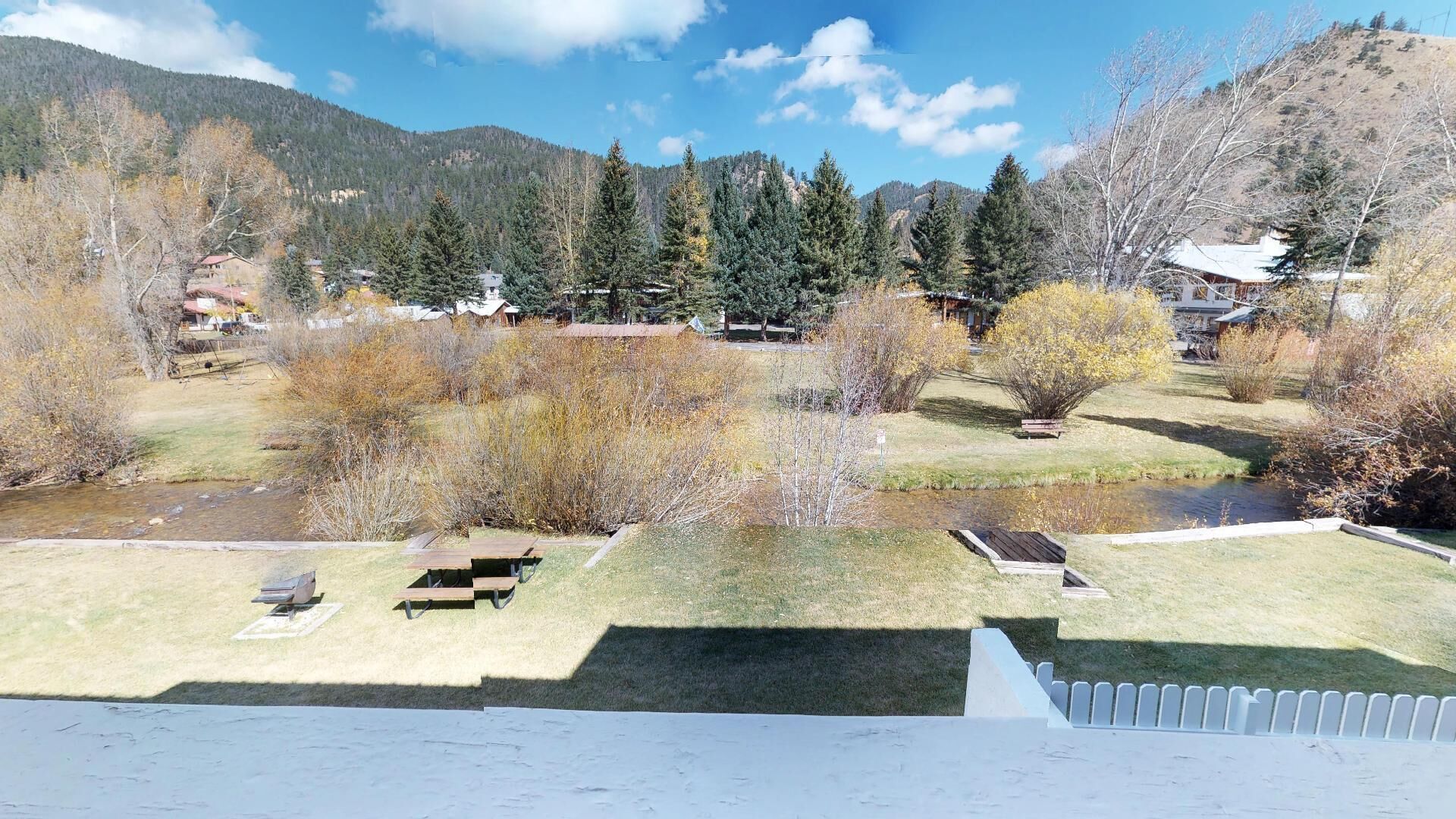 A view of a riverside area with picnic tables, a small bridge, and houses in the background.