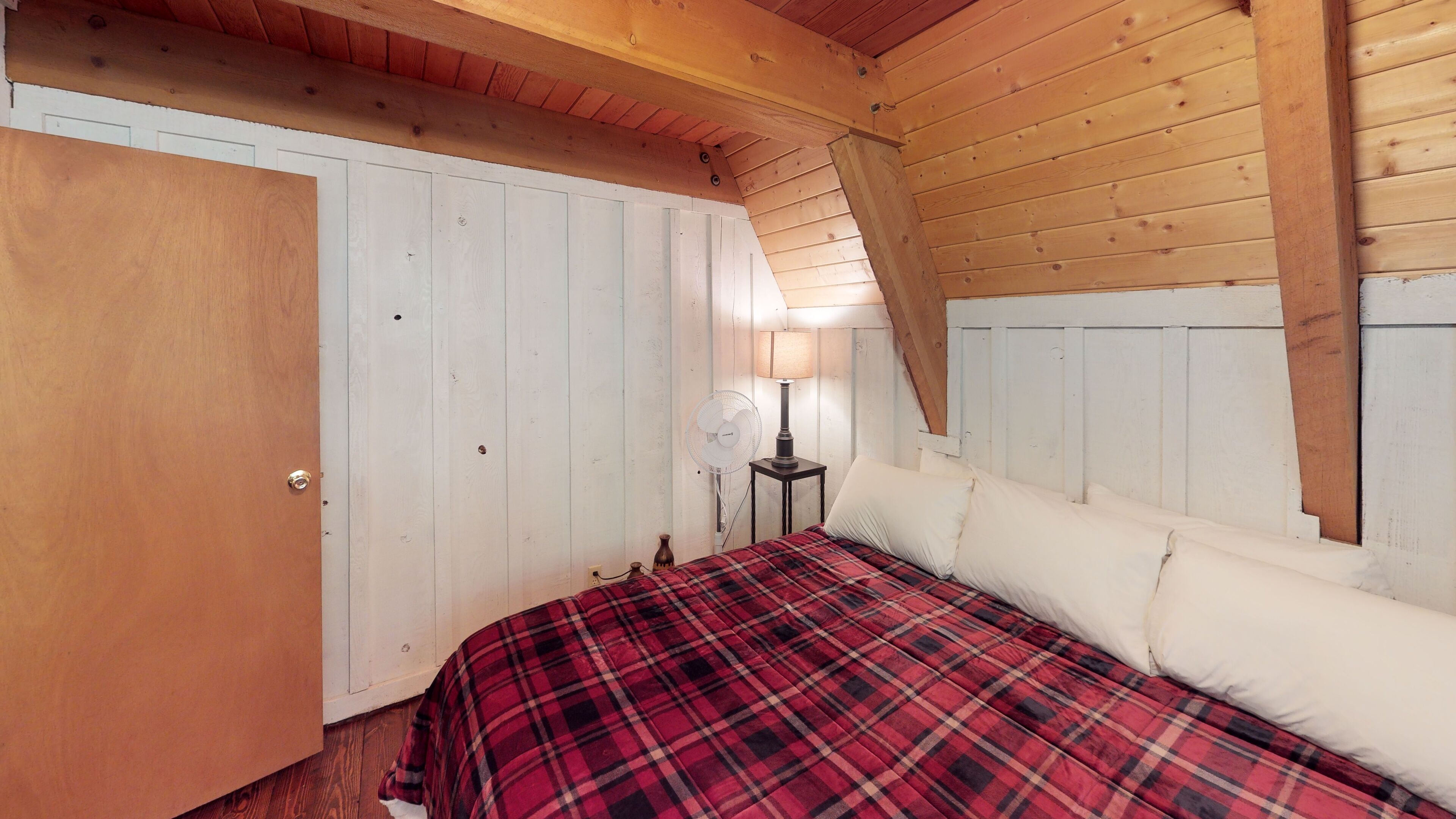 A bedroom with a wooden ceiling, a bed with a plaid blanket, a bedside lamp, and a white door.