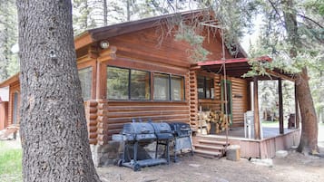 A log cabin with a porch, a grill, and a stack of firewood.