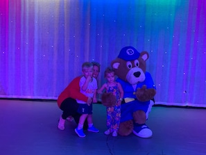 A person in a bear costume with a blue cap and a child in a floral dress posing with a child in a red shirt and black shorts.