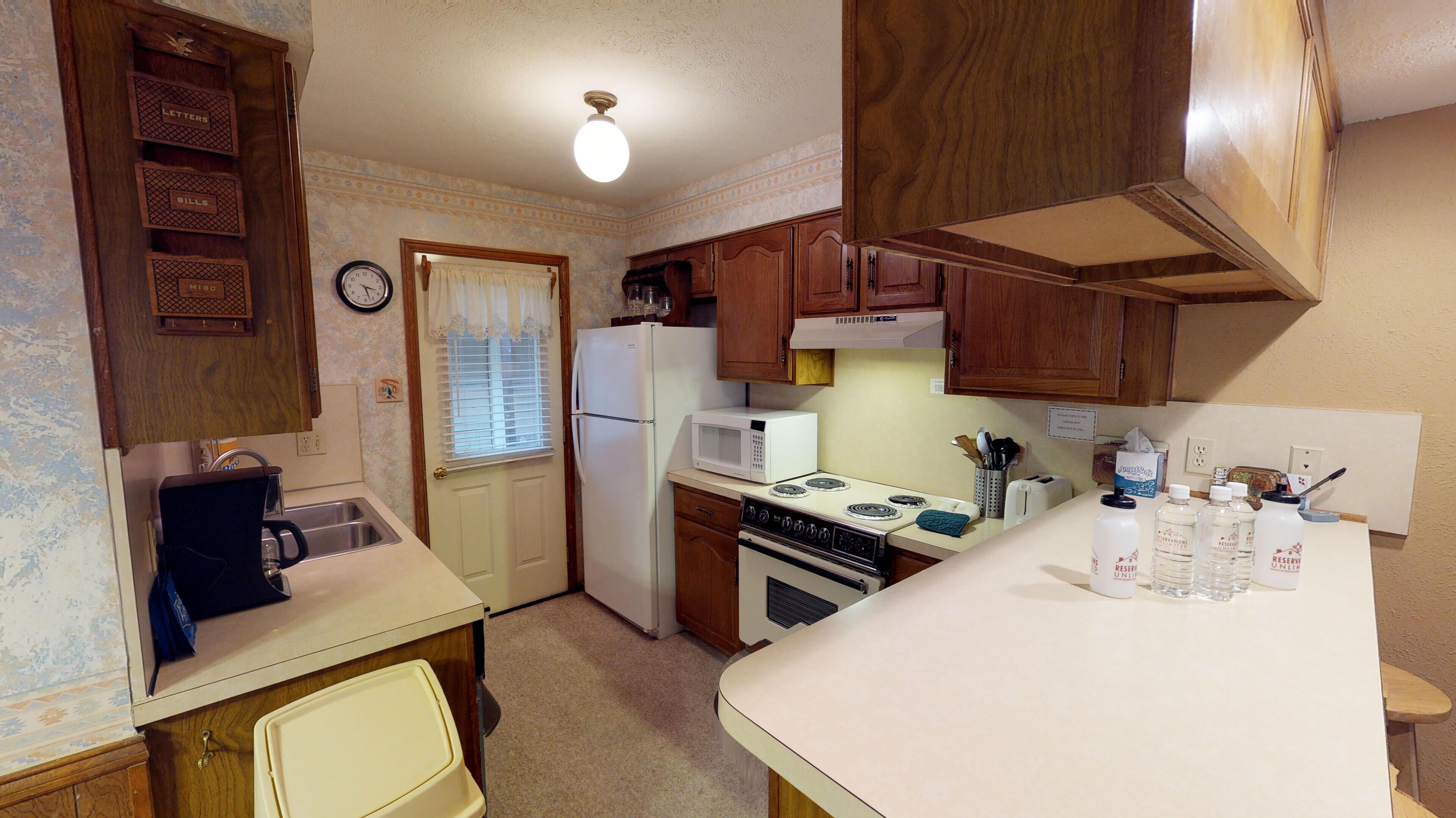 A compact kitchen with a refrigerator, stove, microwave, and sink. There are wooden cabinets and a clock on the wall.