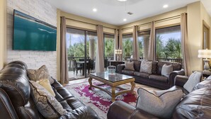 A spacious living room with leather sofas, a glass coffee table, and a large TV mounted on a white brick wall.