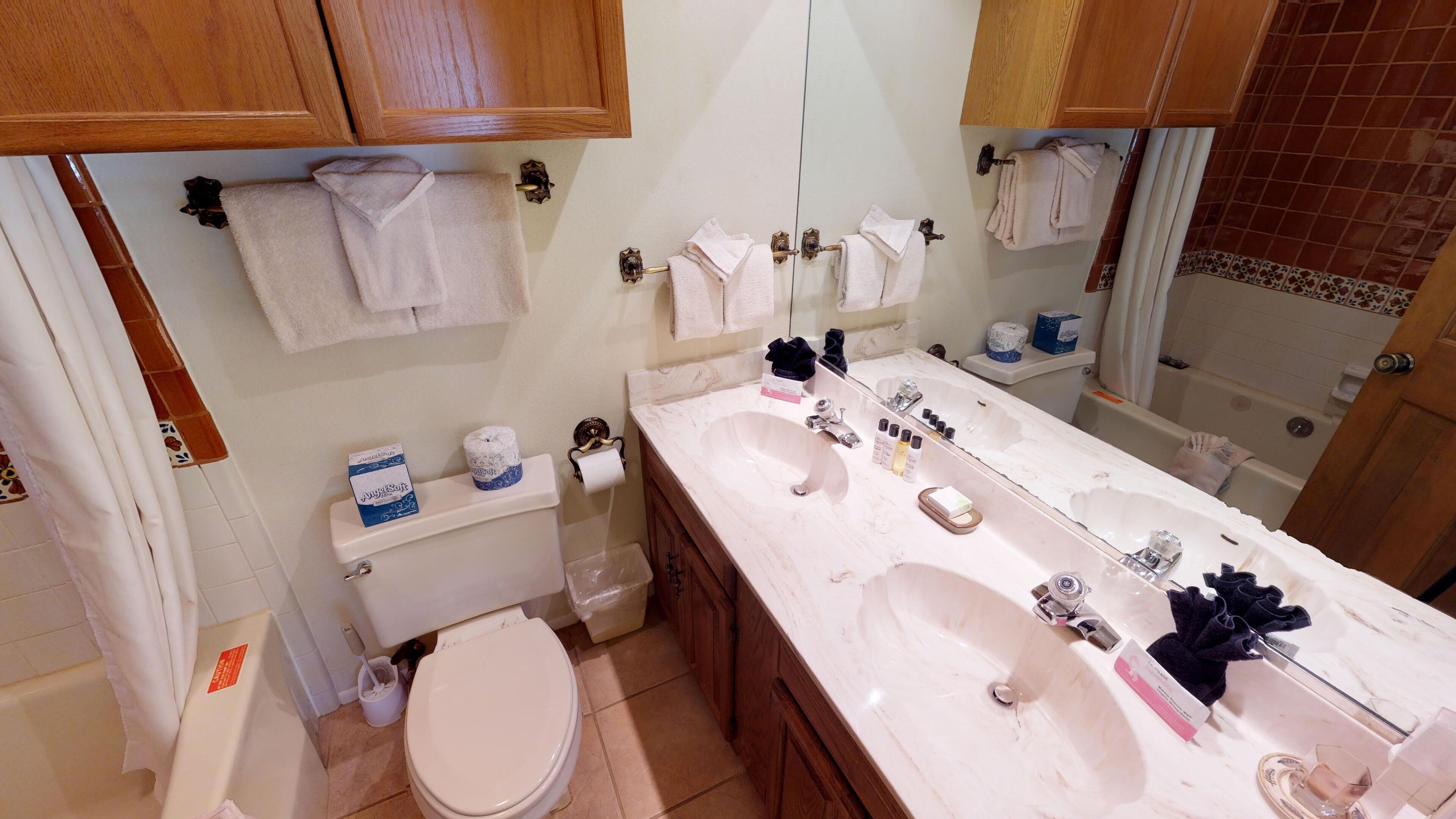 A bathroom with a double sink, toilet, and a mirror. There are towels hanging on the wall and a toilet paper holder.