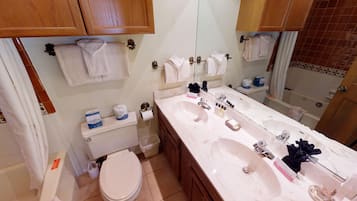 A bathroom with a double sink, toilet, and a mirror. There are towels hanging on the wall and a toilet paper holder.