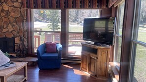 A cozy living room with a stone fireplace, a blue armchair, a television on a wooden stand, and large windows offering a view of a deck and trees. - Valley View in the Upper Valley (Red River)