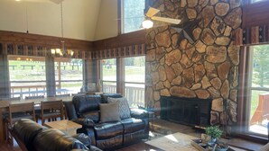 A cozy living room with a stone fireplace, leather sofas, wooden tables, and large windows offering a view of the outdoors. - Valley View in the Upper Valley (Red River)