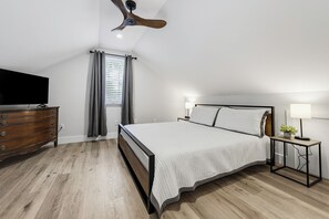 Une chambre à coucher comprenant un lit, une commode, un ventilateur de plafond, une fenêtre avec des rideaux et une table de chevet avec une lampe.