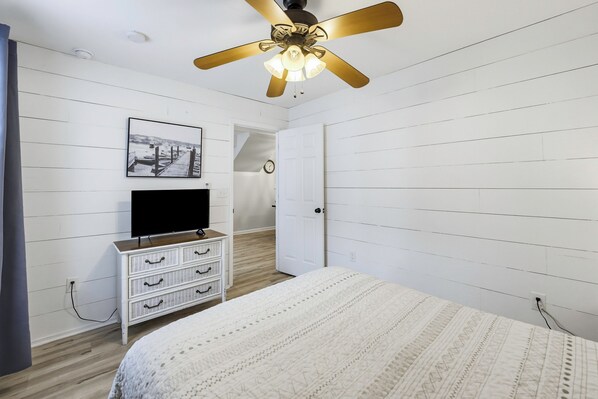 Een slaapkamer met een bed, een dressoir, een plafondventilator, een aan de muur bevestigde televisie en een schilderij.