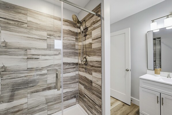 Un baño moderno con ducha envuelta en vidrio, tocador blanco con espejo y puerta que da a otra habitación.