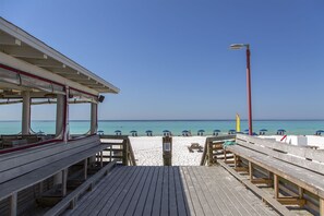 A wooden pier with benches and umbrellas, leading to a beach with clear blue water and a few people in the distance.