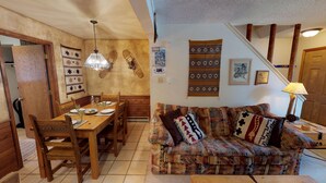 A cozy living room with a wooden dining table set for four, a patterned sofa, and a staircase with wooden railings.