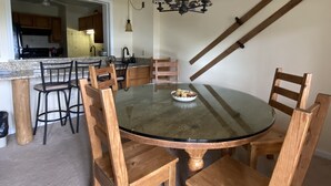 Un'area pranzo con un tavolo rotondo in vetro circondato da sedie di legno, un lampadario e una cucina sullo sfondo.