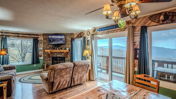 A cozy living room with a fireplace, a large window overlooking a deck, and a wooden dining table. - Sugar Ski Premium Two Bedroom - Unit 821 (Sugar Mountain)