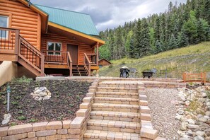 Une cabane en rondins avec un toit végétalisé, une terrasse en bois et un escalier en pierre menant à un espace patio aménagé avec des sièges d’extérieur et un barbecue.