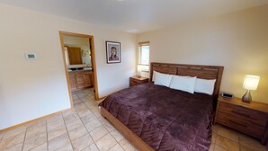 A bedroom with a large bed, wooden headboard, bedside tables with lamps, a nightstand with a clock, and a bathroom visible through an open door. - 29 Valley of the Pines with Hot Tub in the Upper Valley (Red River)
