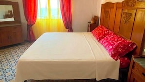 Un dormitorio con cama, cortinas, cabecera de madera, un cuadro, cómoda y suelo estampado.