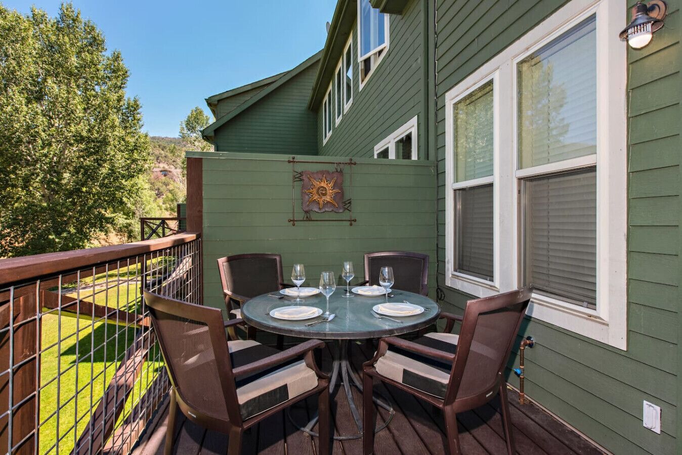 A deck with a round table set for four, surrounded by chairs, against a green house wall.
