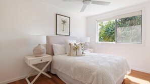Une chambre à coucher comprenant un lit, des tables de chevet, un ventilateur de plafond, une fenêtre donnant sur un paysage verdoyant et un tableau encadré accroché au mur.