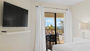 Habitación de hotel con televisor montado en la pared, balcón con mesa y sillas, y vista al océano.