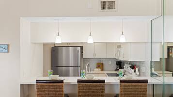 A modern kitchen with a stainless steel refrigerator, white cabinets, and a glass island with wicker chairs.