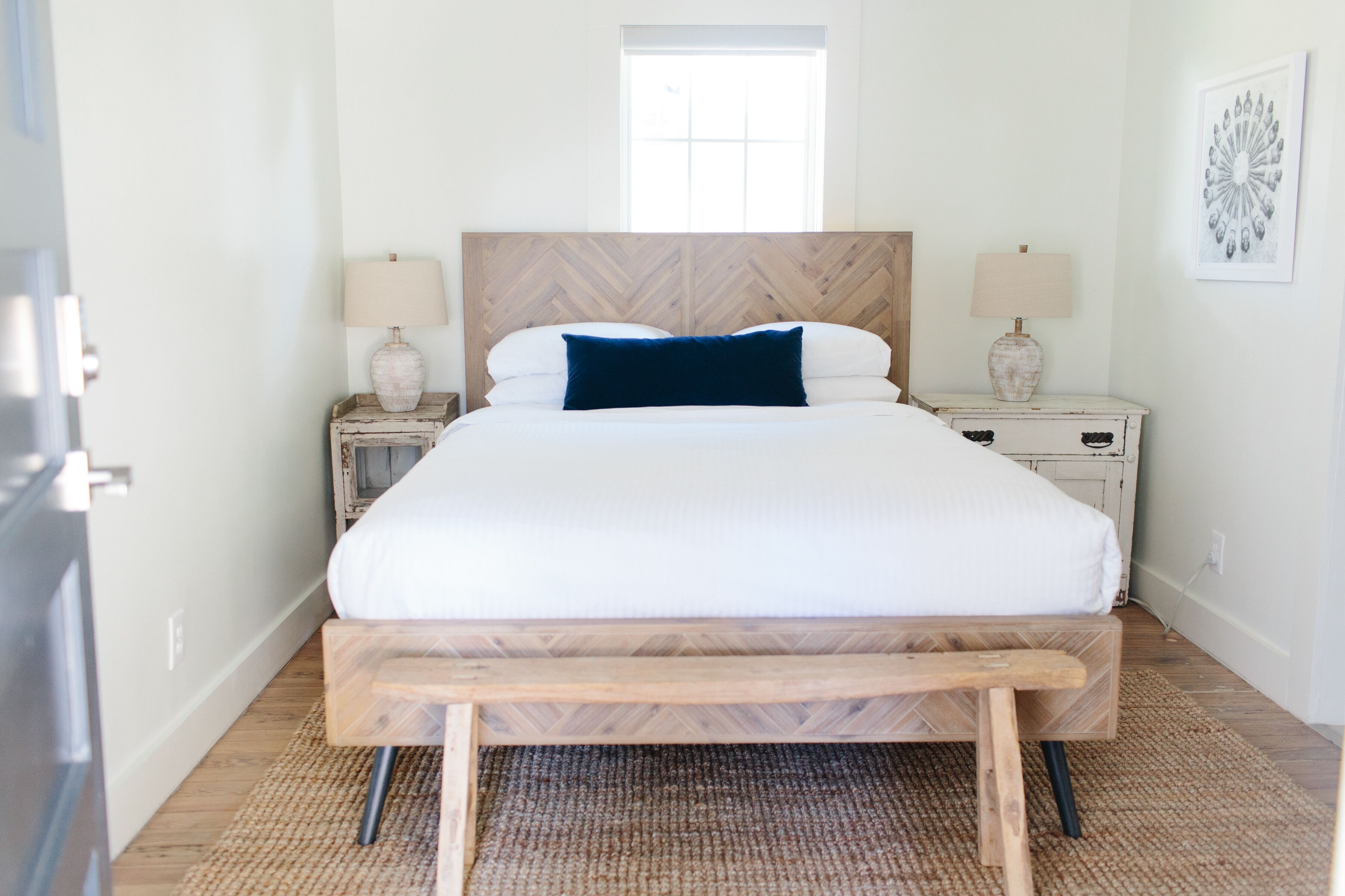 Una cama grande con cabecera de madera, dos mesitas de noche con lámparas, un banco al pie de la cama y una ventana sobre ella.