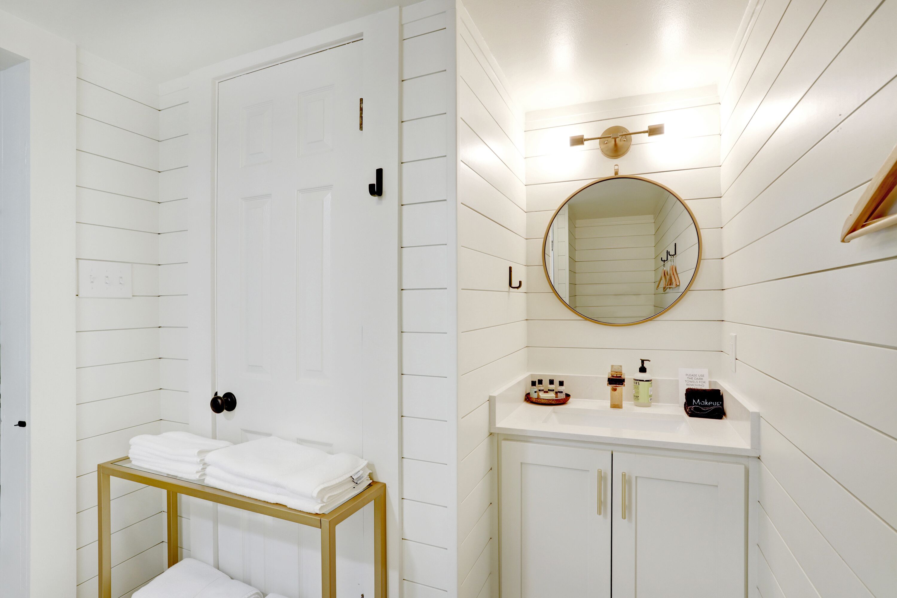 A bathroom with a white door, a wooden-framed shelf with towels, a vanity with a mirror, and a sink.