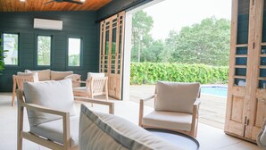 Zona d'estar interior acollidora amb cadires i un sofà de fusta, un pont de fusta que condueix a una piscina i vista a la vegetació exterior.