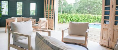 Un área de estar interior acogedora con sillas y un sofá de madera, una terraza de madera que da a una piscina, y vistas de follaje al exterior.