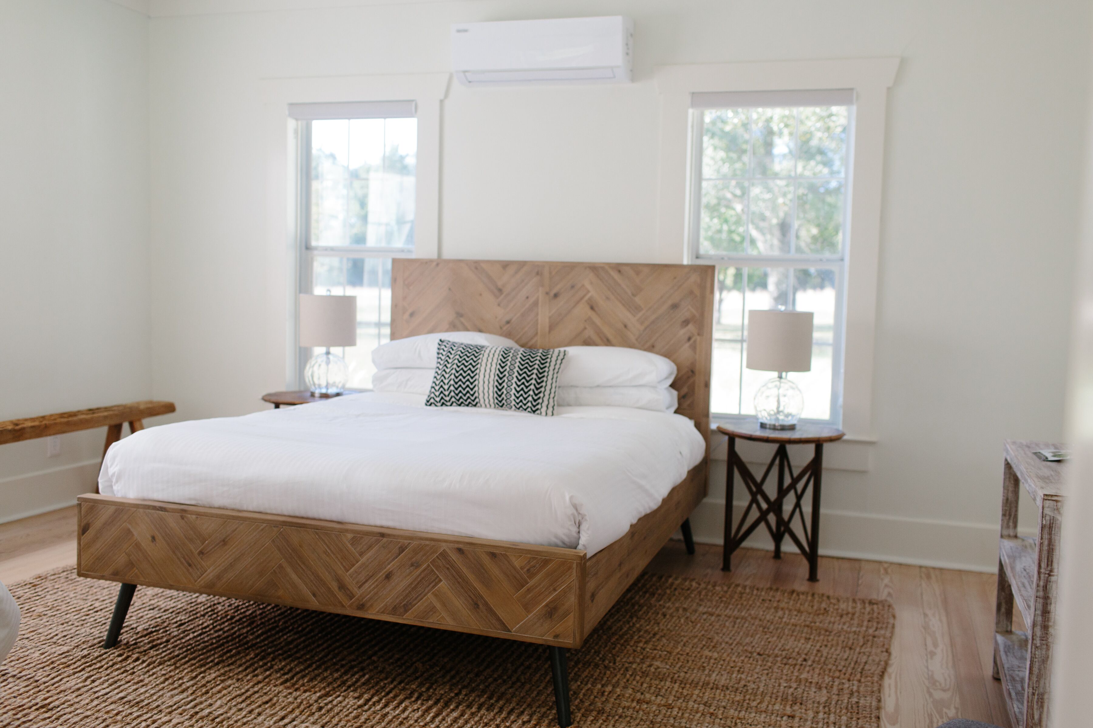 A bedroom with a wooden herringbone bed frame, white bedding, two bedside lamps, and a large window providing natural light.