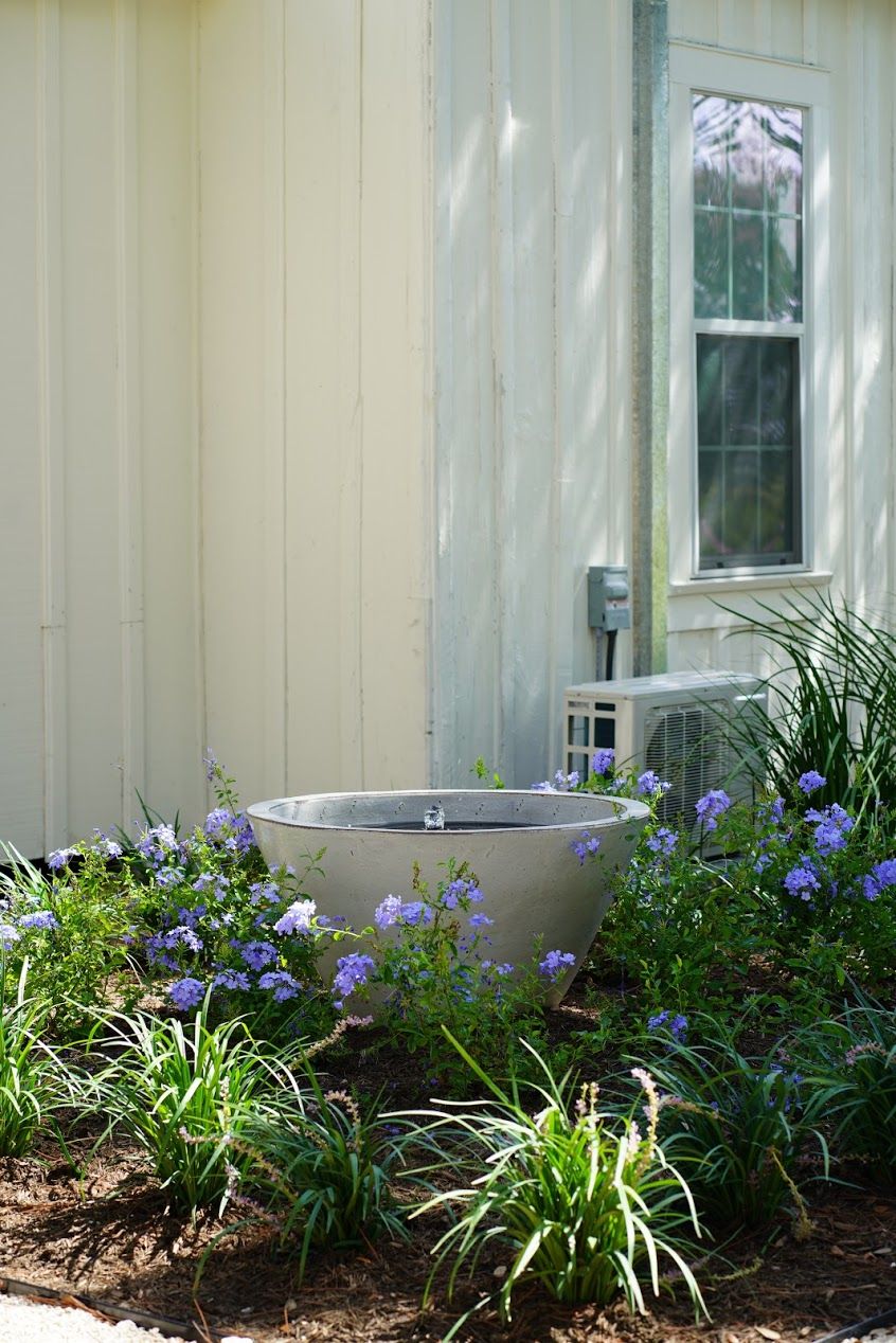 A house with a large metal water feature in the garden, surrounded by purple flowers and green plants.