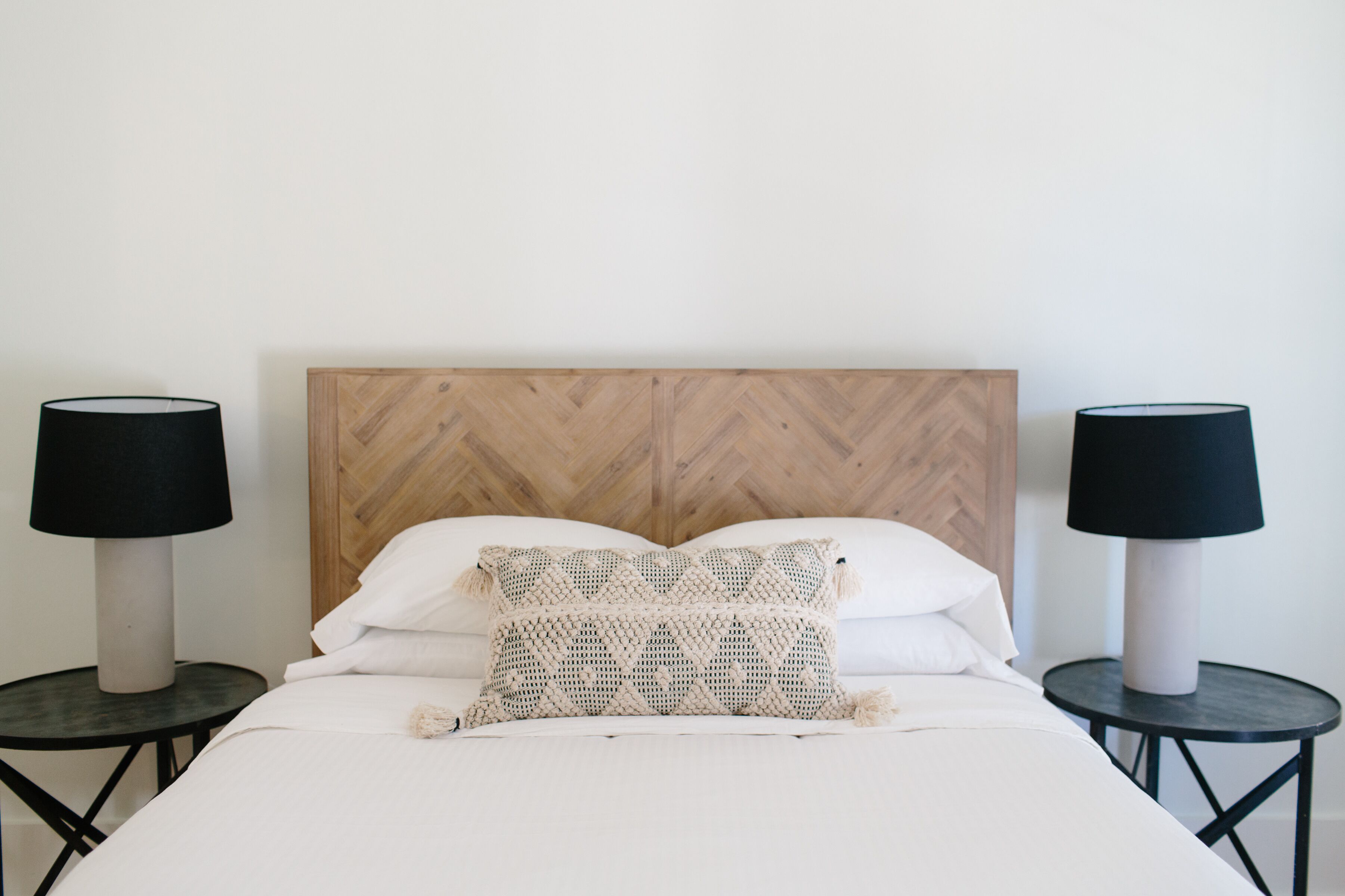 Una cama bien hecha con cabecera de madera, dos mesitas de noche con lámparas y un cojín decorativo.