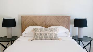 Una cama bien hecha con cabecera de madera, dos mesitas de noche con lámparas y un cojín decorativo.