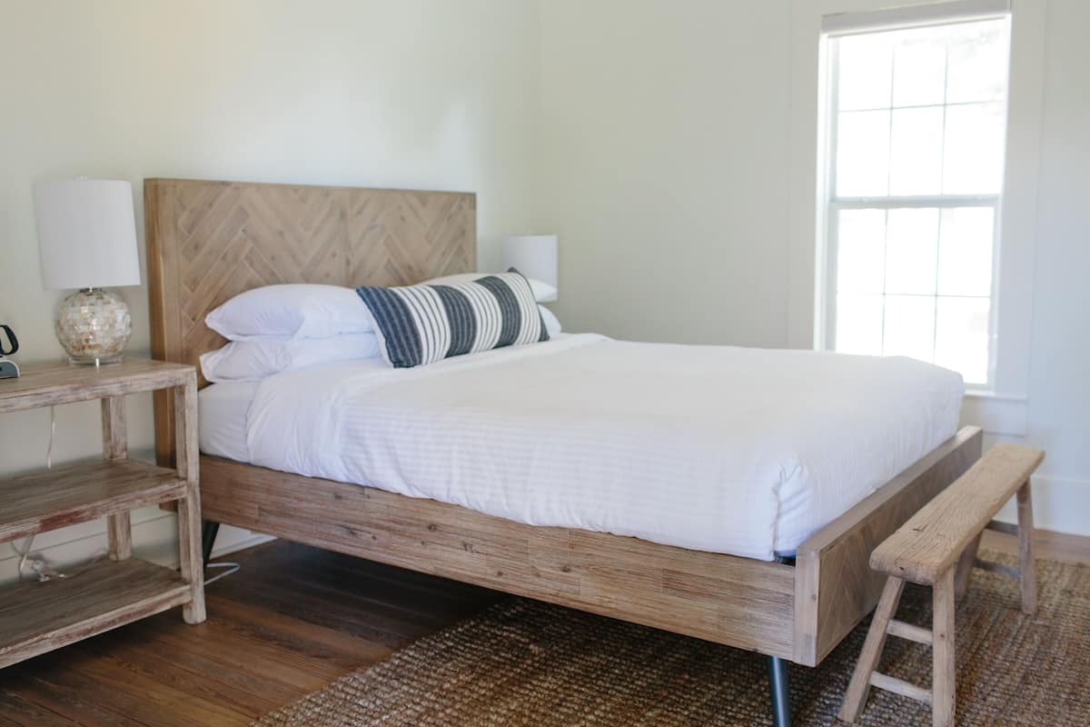 Ein ordentlich bezogenes Bett mit einem hölzernen Kopfteil, einem Nachttisch mit Lampe und einer Bank am Fußende.
